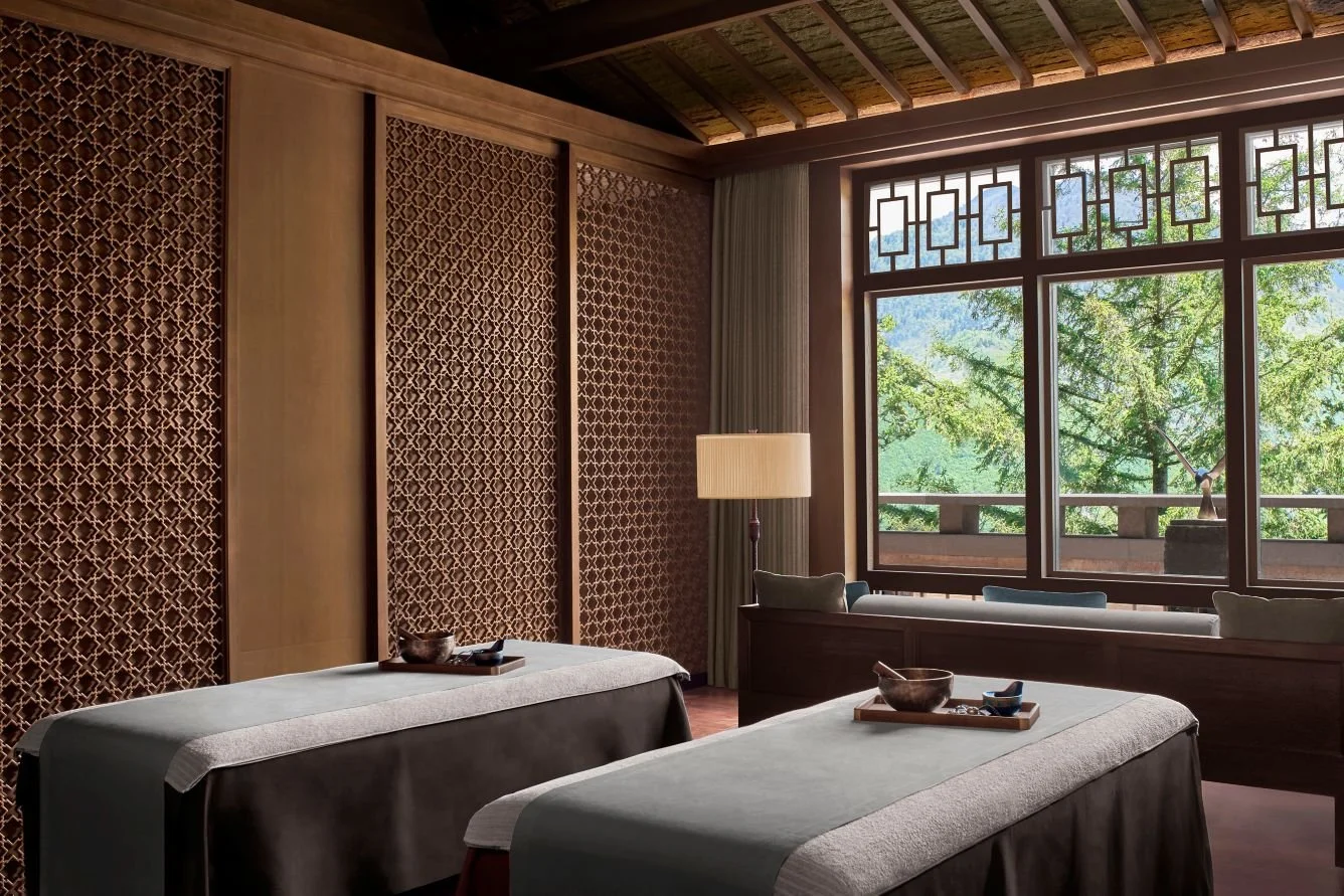 Spa treatment rooms with two massage beds, wooden walls, large window with a view of green trees, and a lamp.