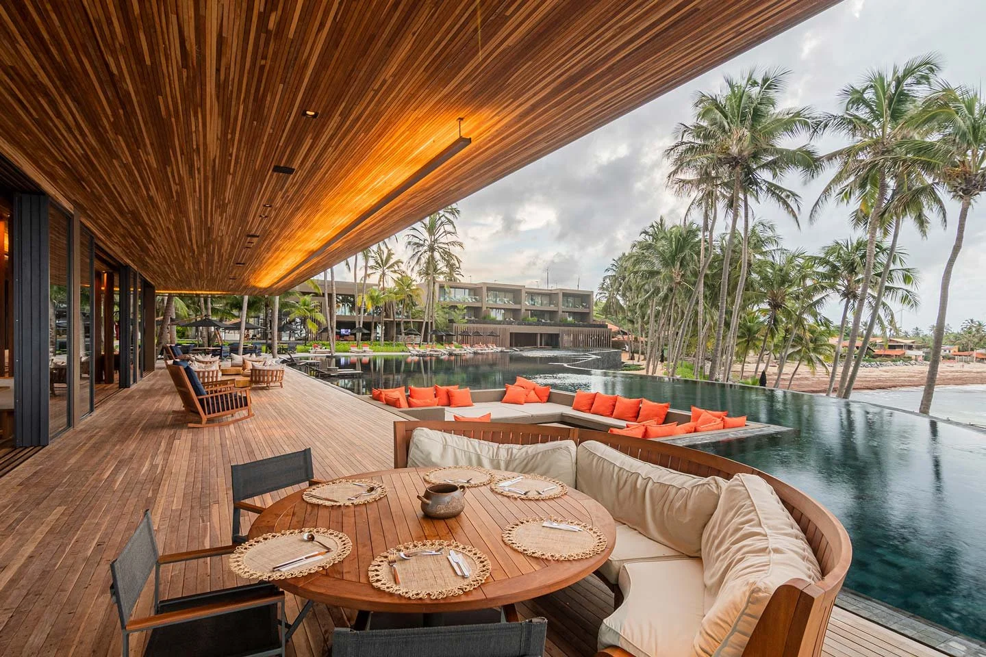 Outdoor wooden deck with a dining table and chairs, a built-in sofa with orange cushions, a pool with an overhang, and palm trees overlooking the ocean.