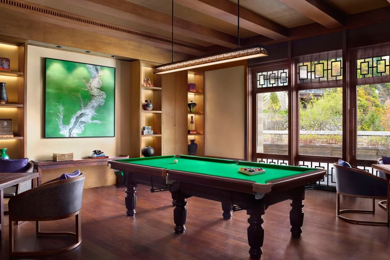A room with a billiards table, wooden floors, shelves with decorative items, a large painting on the wall, and windows showing trees outside.