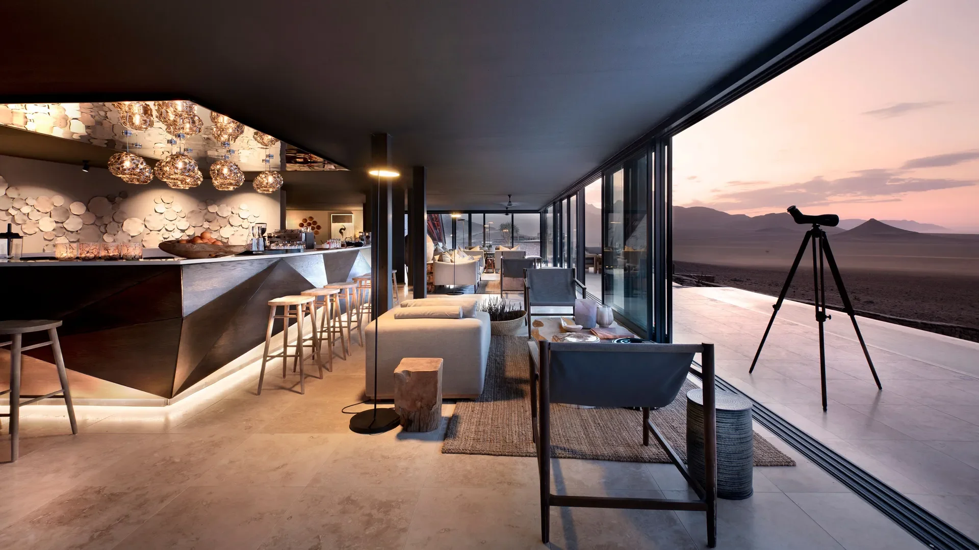 Modern lounge area with large floor-to-ceiling windows overlooking a desert landscape at sunset, a telescope on a tripod aimed outside, and a bar counter with barstools and hanging pendant lights.