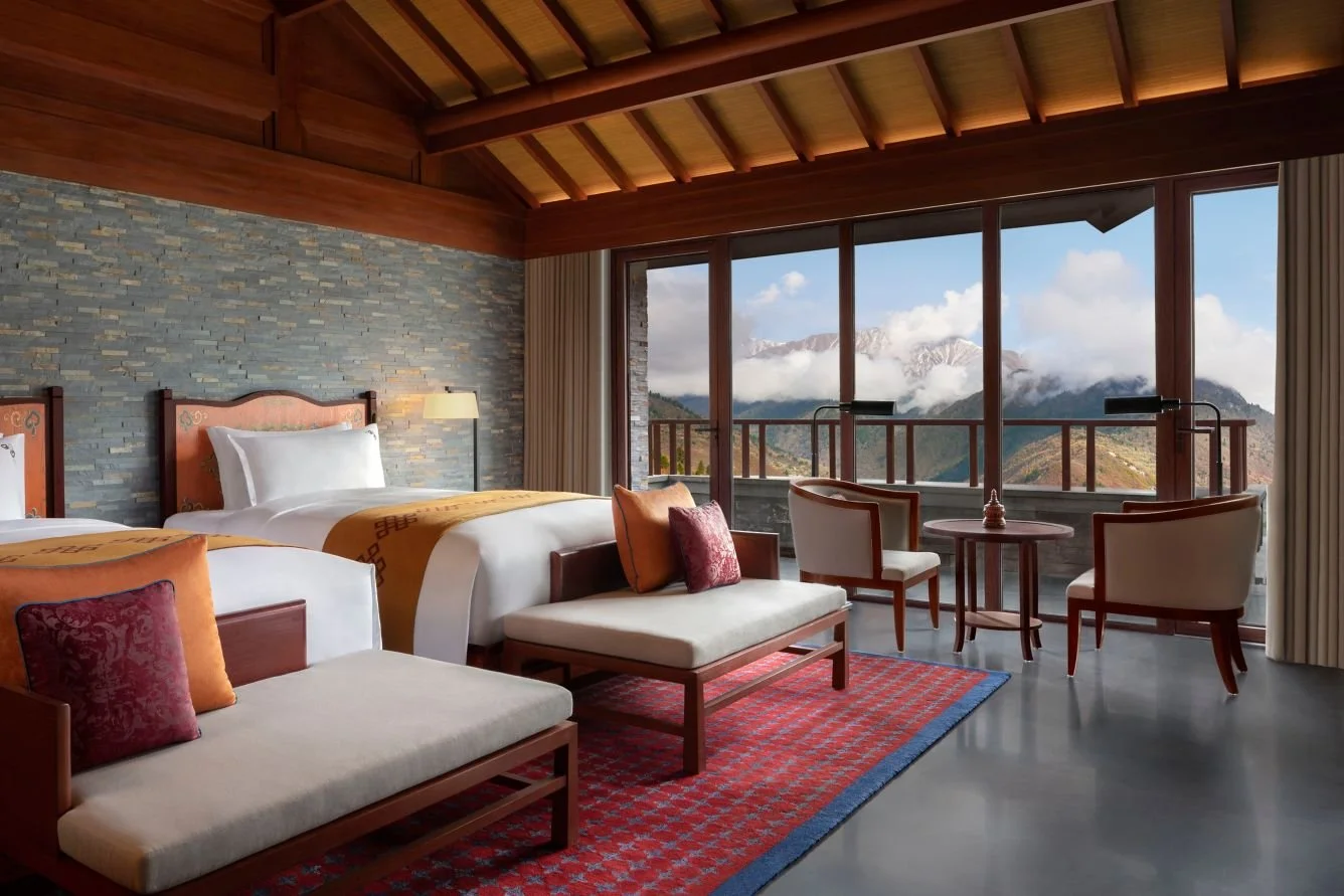 A luxurious hotel room with two beds, a sitting area with chairs and a coffee table, and floor-to-ceiling windows showing mountain scenery with clouds and snow-capped peaks.