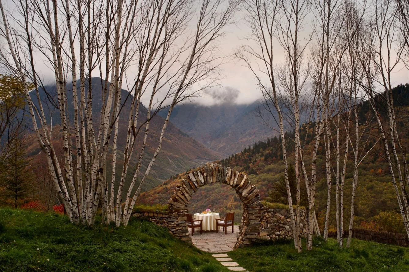 A scenic outdoor setting with a stone archway framing a table with a white tablecloth and two chairs, surrounded by leafless birch trees, mountainous landscape with clouds in the background.