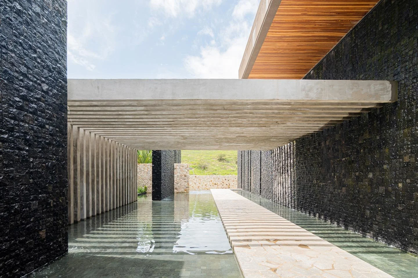 Modern architectural structure with water feature, black and beige stone walls, wooden ceiling detail, and pathways leading outward, under a partly cloudy sky.