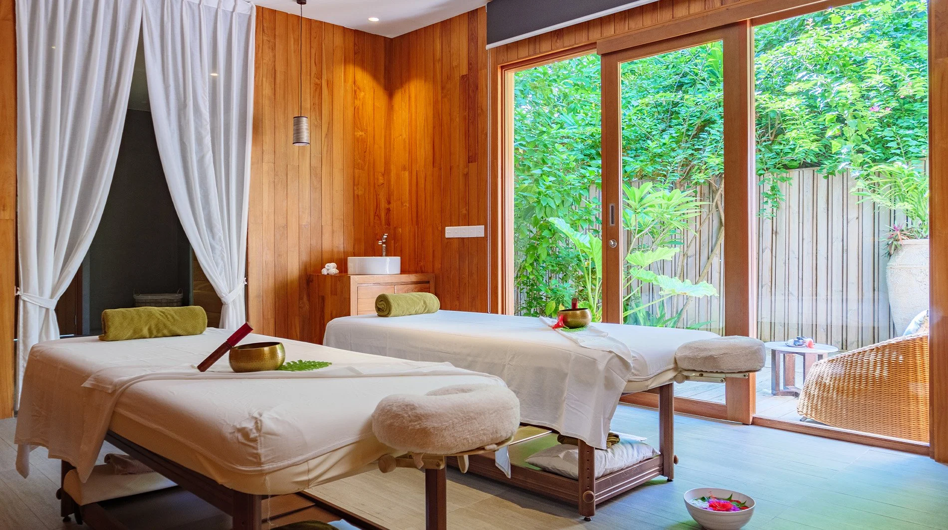 Two massage tables in a spa room with wooden walls, large sliding glass door, and lush greenery outside, with towels, bowls, and soothing decor.