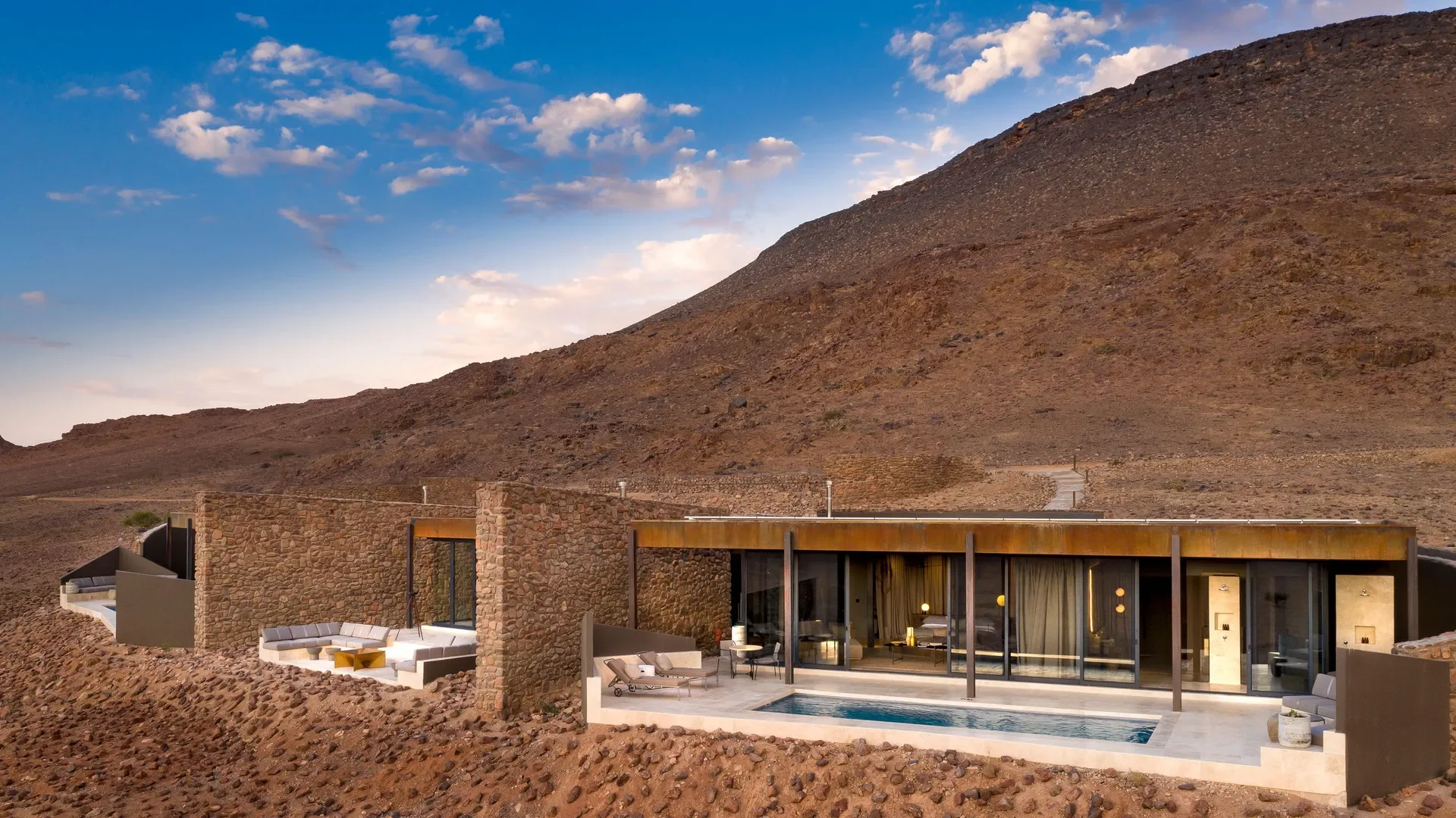 Modern house with stone and glass exterior, outdoor pool, and patio furniture, situated on arid, rocky terrain with hills in the background and a partly cloudy sky.
