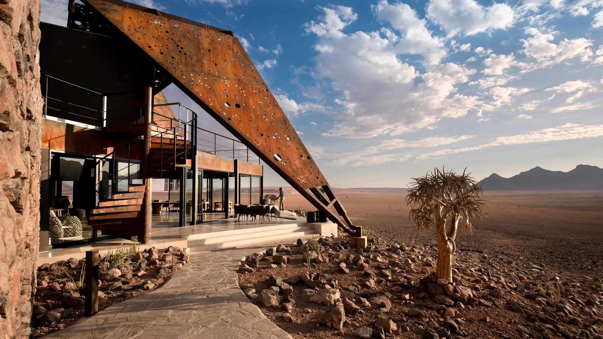 Modern house with rust-colored curved metal roof and large glass windows set in a desert landscape with distant mountains, a lone tree, and a person outside.