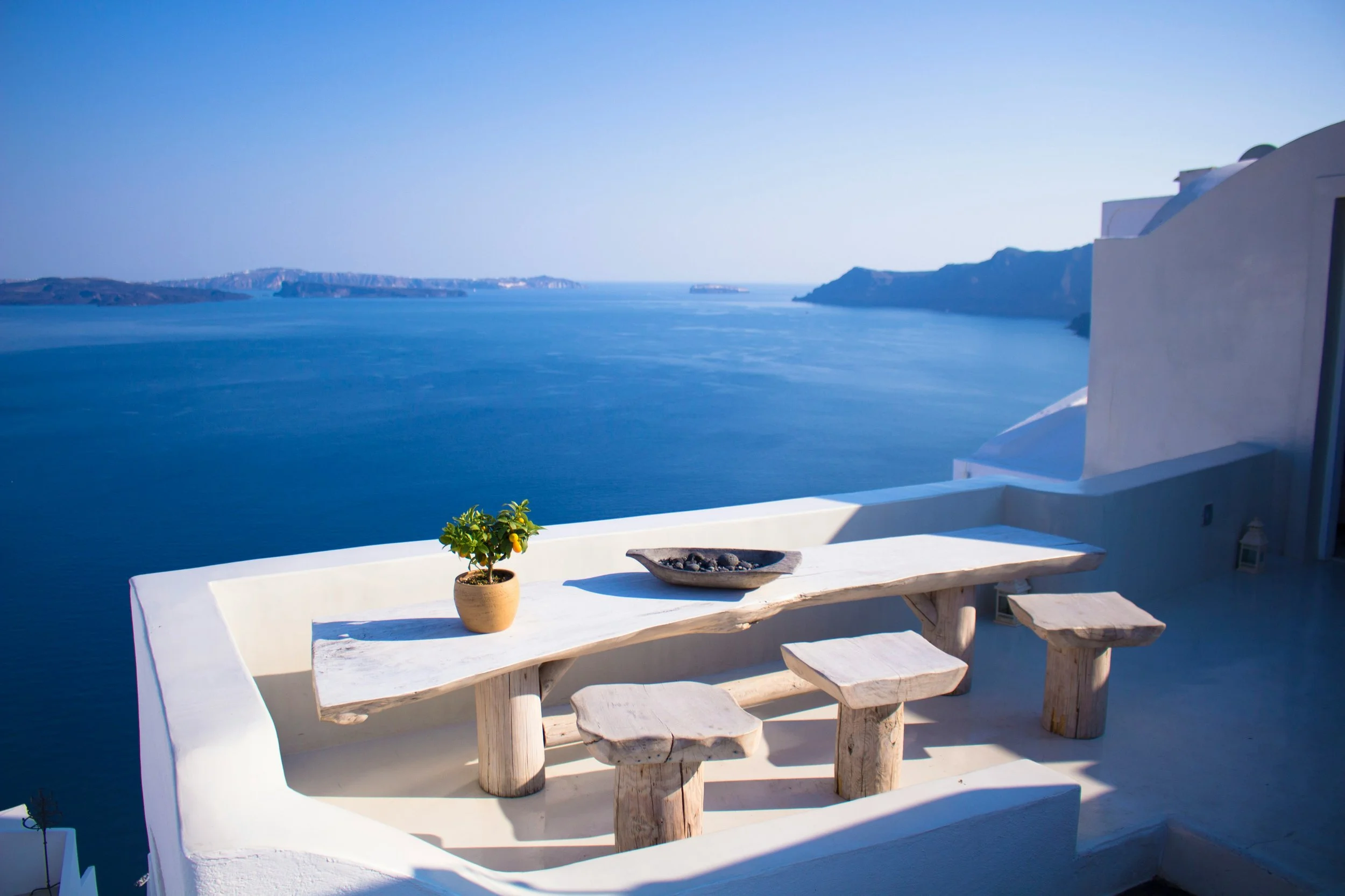 A balcony with a white wooden table and four stools, a small potted lemon tree, and a shallow bowl with stones, overlooking a calm blue sea and distant islands on a sunny day.