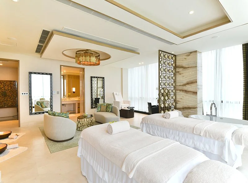 Luxurious hotel room with two massage beds, seating area, large windows with sheer curtains, and modern decor.