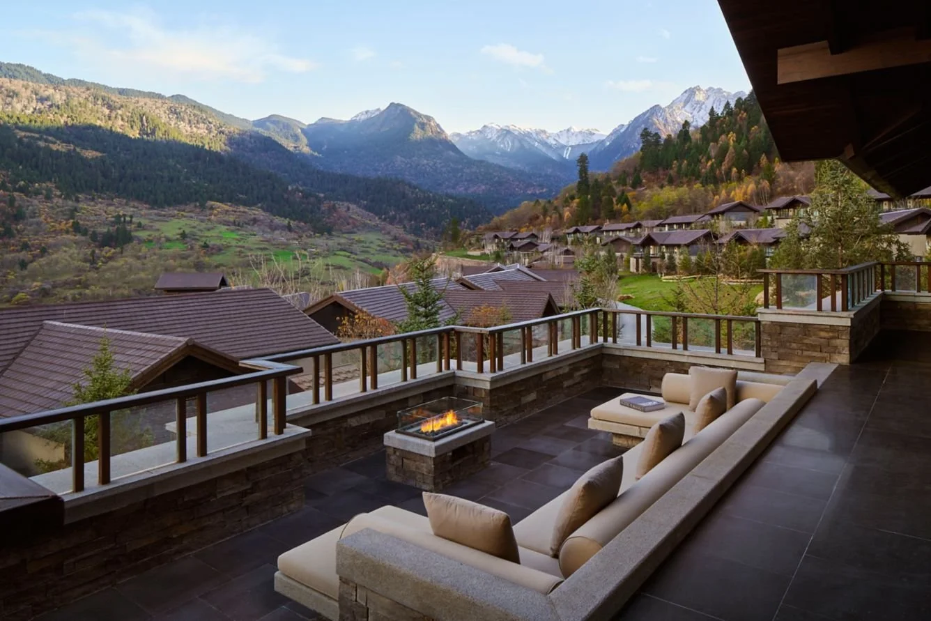 A spacious balcony with cream-colored cushioned seating, a small fire pit, and a mountain view with houses, trees, and snow-capped peaks.