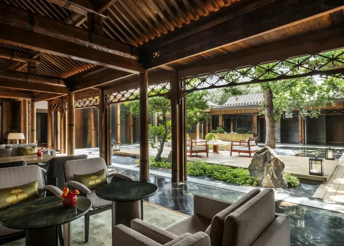 Indoor lounge area with beige armchairs and black coffee tables, open view of an outdoor garden with trees, stone, wooden benches, and lanterns, in traditional Asian style.
