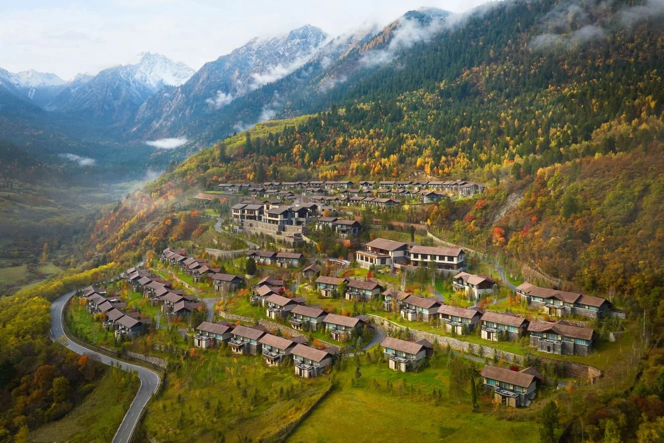 Aerial view of a hillside residential area with houses, surrounded by autumn foliage and mountains in the background.