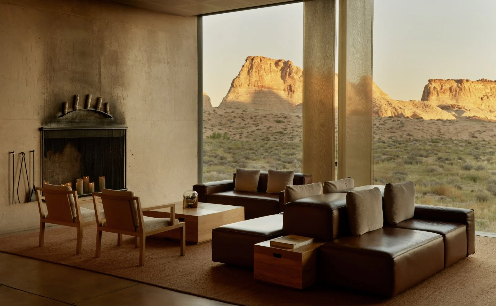 Modern living room with a large window overlooking desert landscape with rock formations in the distance, featuring leather sofas, wooden armchairs, a fireplace with candles, and minimalist decor.