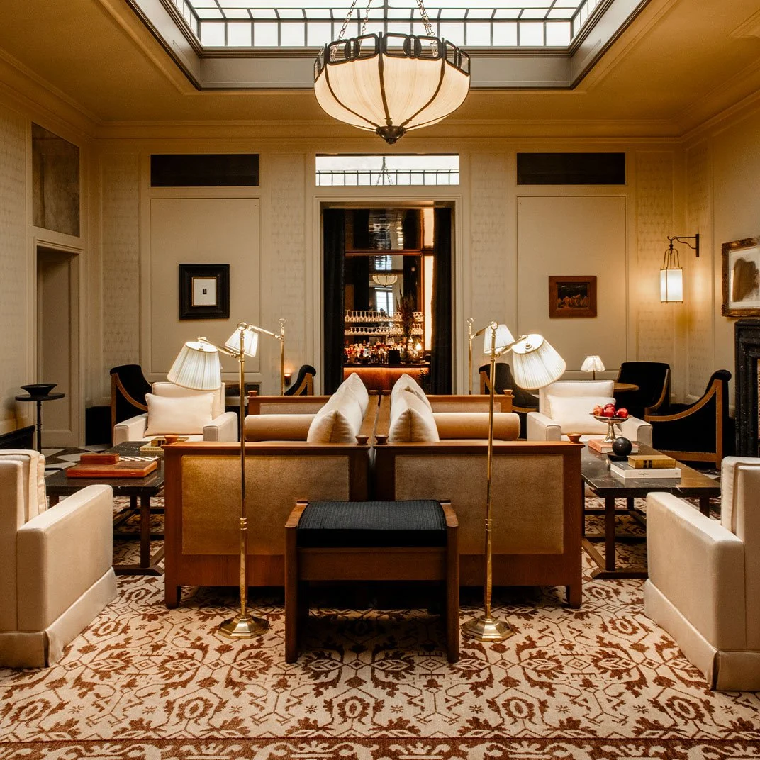 Elegant hotel lounge with beige sofas, black armchairs, and wooden coffee tables, featuring a patterned rug, large chandelier, and a skylight ceiling.