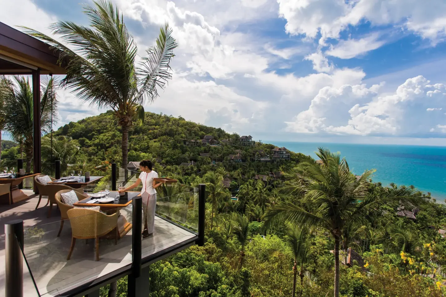 A terrace with a woman setting a table overlooking a lush green hillside and the ocean under a partly cloudy sky.