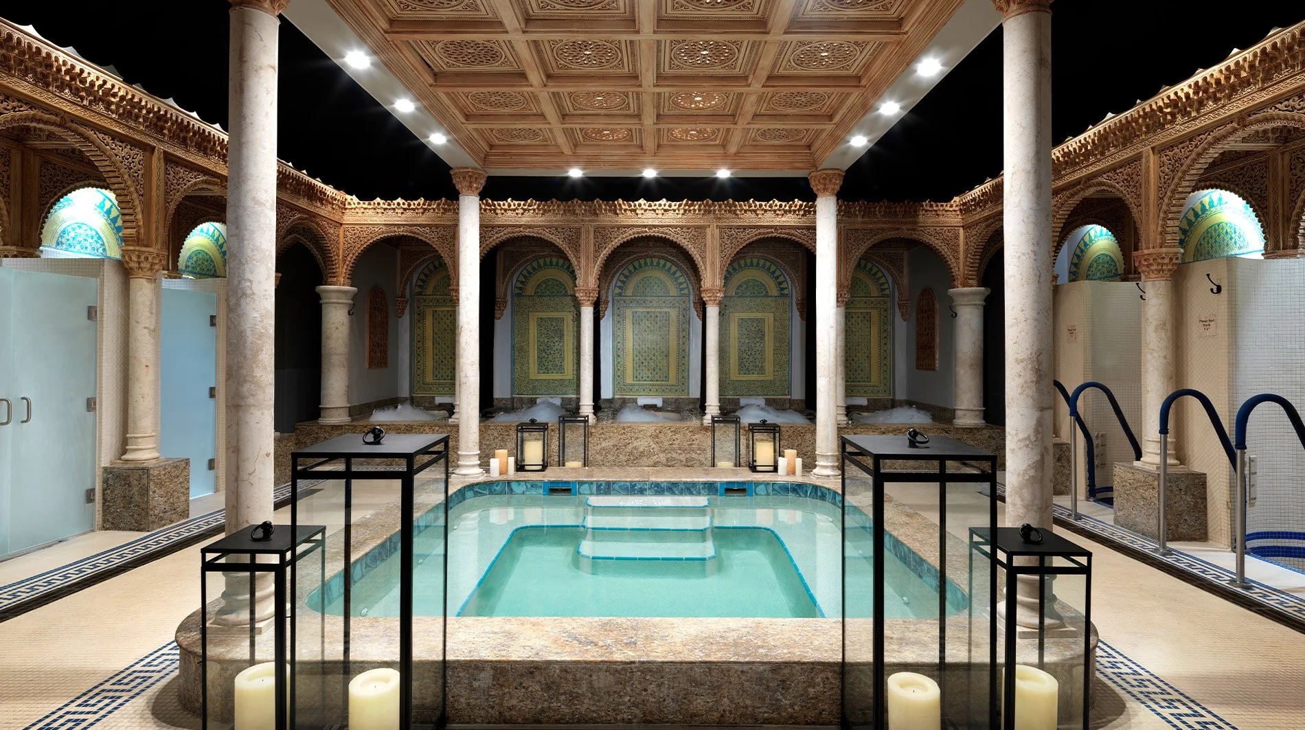 Indoor luxurious spa with ornate wooden decor, columns, and a small pool with steps, candles, and lanterns.