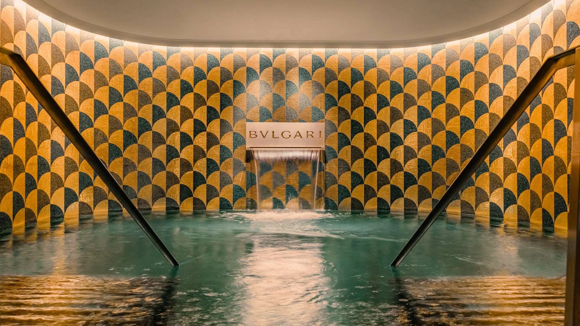 Indoor spa with a waterfall feature and patterned tile walls, featuring the BVLGARI logo.