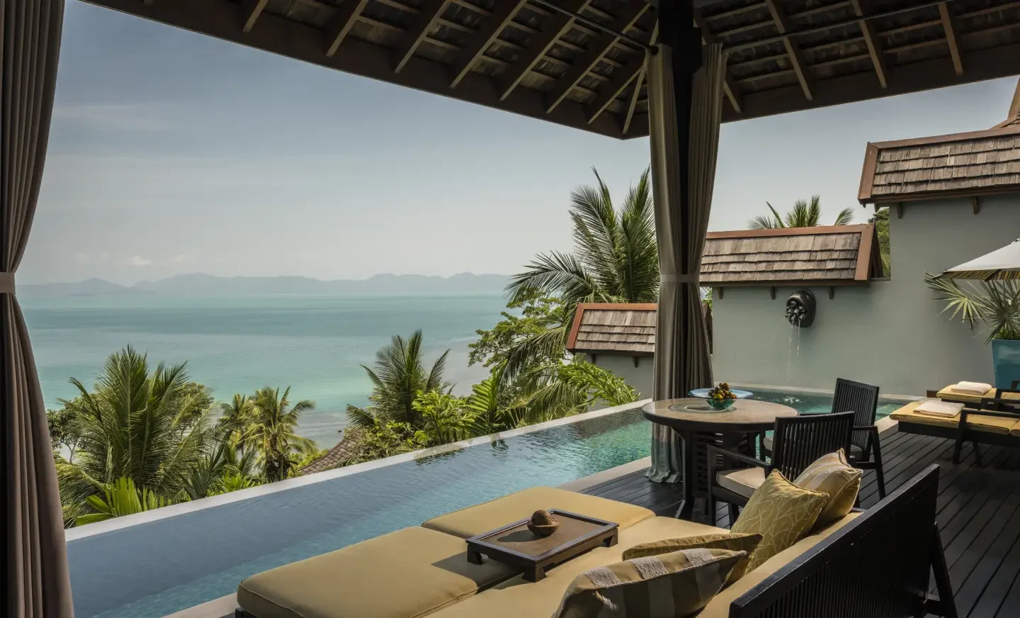 A view from a balcony overlooking tropical trees, water, and distant islands. The balcony features an infinity pool, outdoor seating, and a shaded area with curtains.