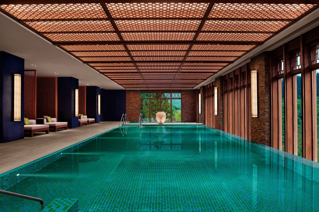 Indoor swimming pool with large windows, wooden walls, and cushioned seating area along the side.