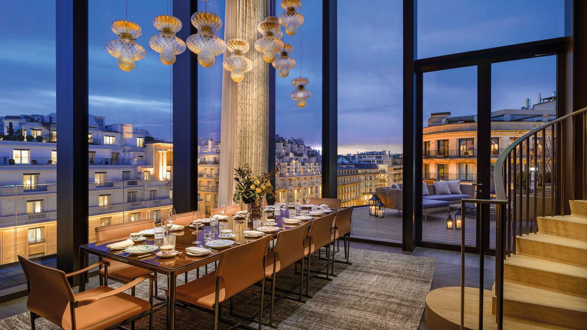 Elegant dining room with a view of city buildings at dusk, set with a large table, chairs, and a vase with flowers, with a spacious balcony outside.