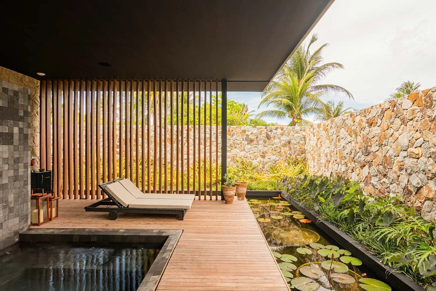 Outdoor patio with lounge chair, small water pond with lily pads, potted plants, stone wall, and palm trees in the background.