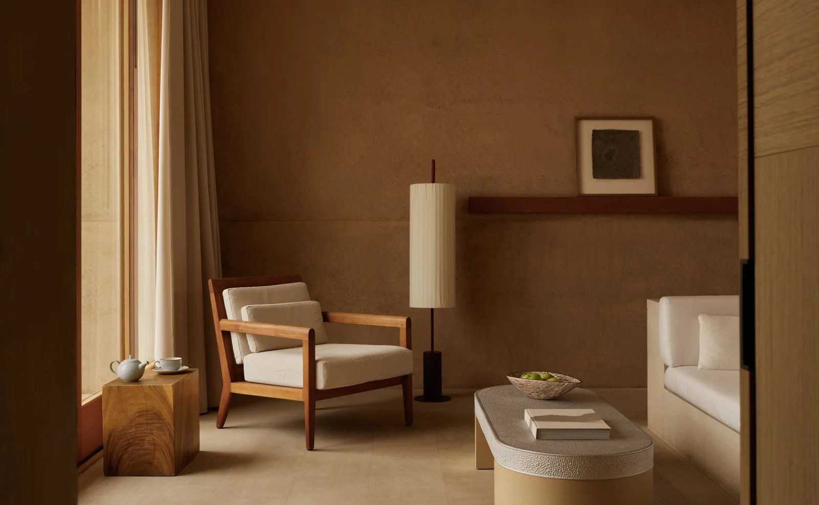 Minimalist living room with beige walls, a window with curtains, a wooden side table with a teapot and a teacup, a cream armchair with wooden armrests, a tall white floor lamp, a framed artwork on a floating shelf, and a beige sofa with a cushion. A 