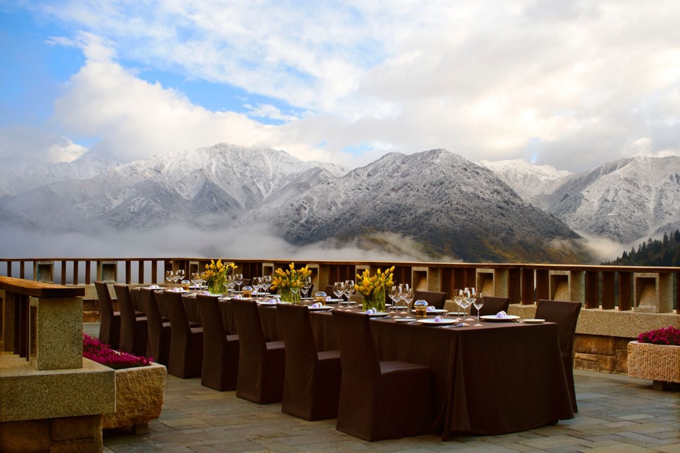 Dining table set with flowers and glassware on a balcony overlooking snow-capped mountains and cloudy sky.
