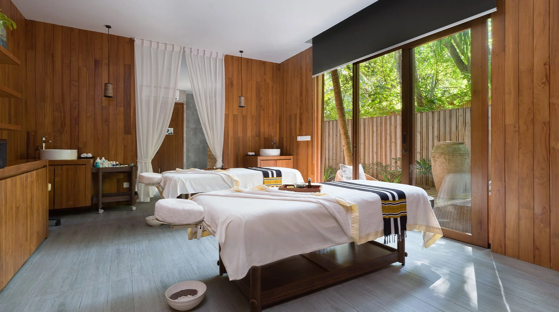 A wellness spa room with two treatment beds covered with white sheets and black-and-yellow striped towels, wooden walls, a sliding glass door leading to a garden with trees and large pottery, and various small spa products on carts and shelves.
