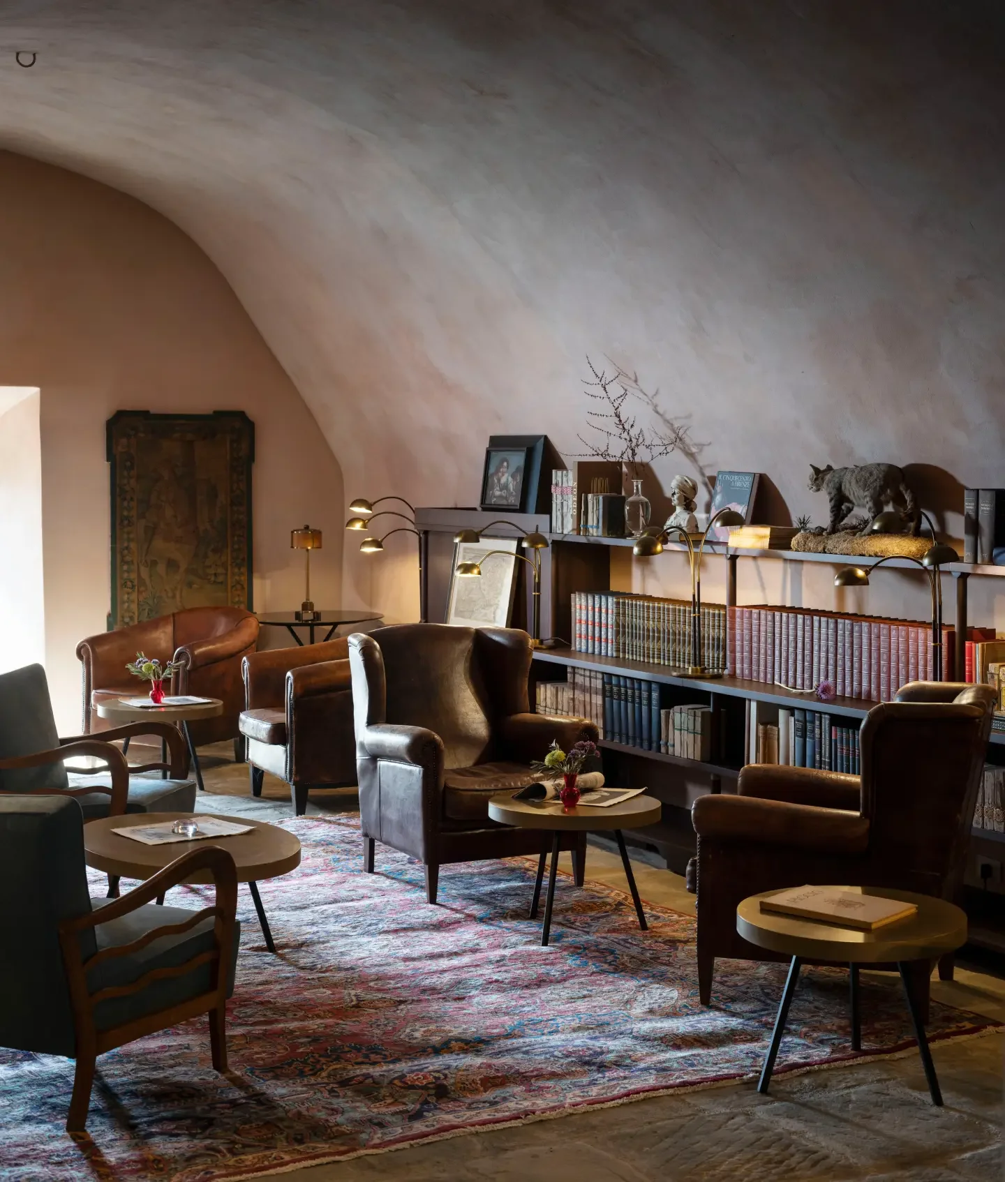 Cozy reading room with leather armchairs, small tables, a bookshelf with decorations including a cat figurine, framed photos, and a tapestry, under a curved ceiling with warm lighting.