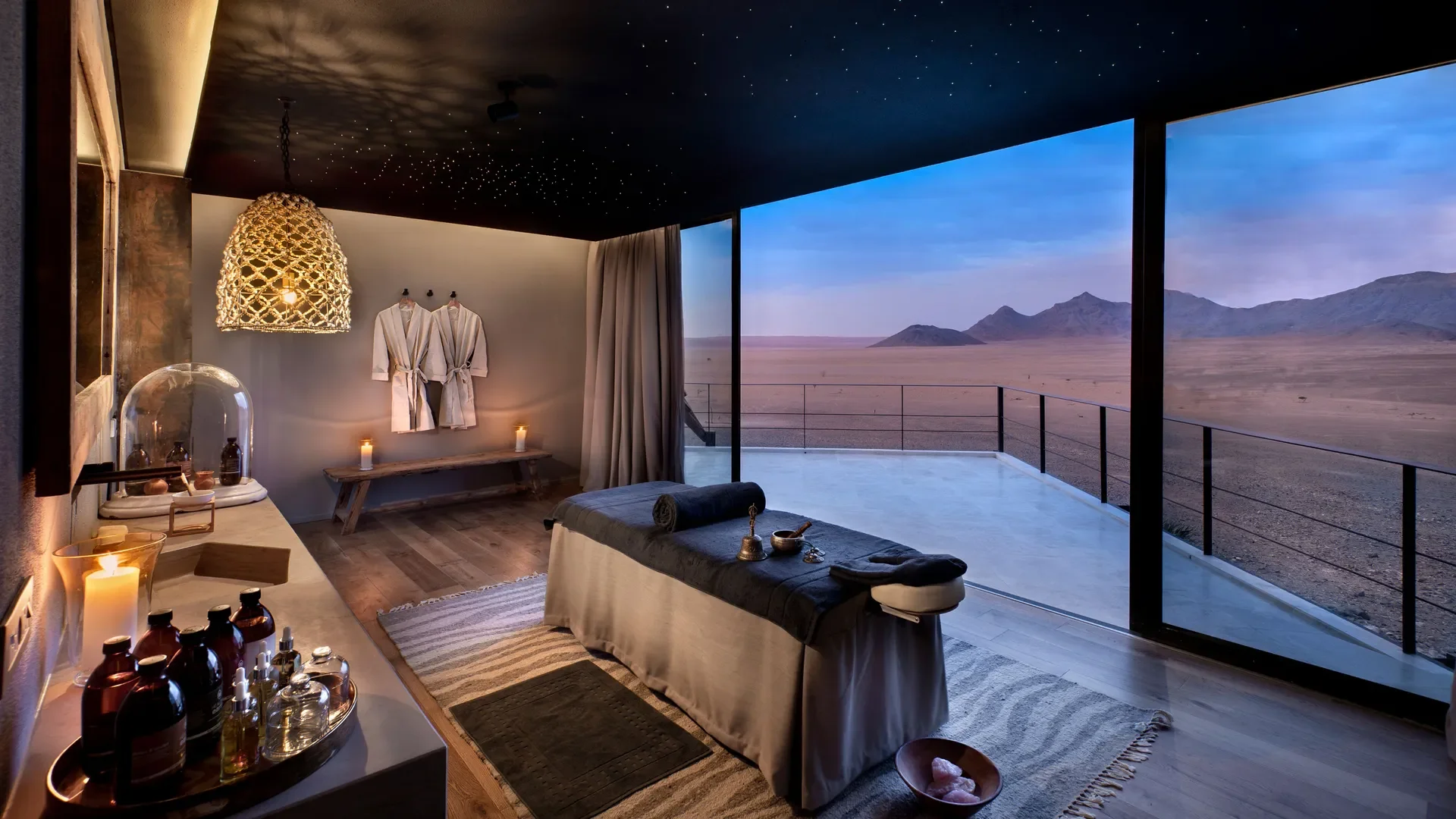 A luxurious spa room with a massage bed, candles, and a scenic desert view through large glass windows.