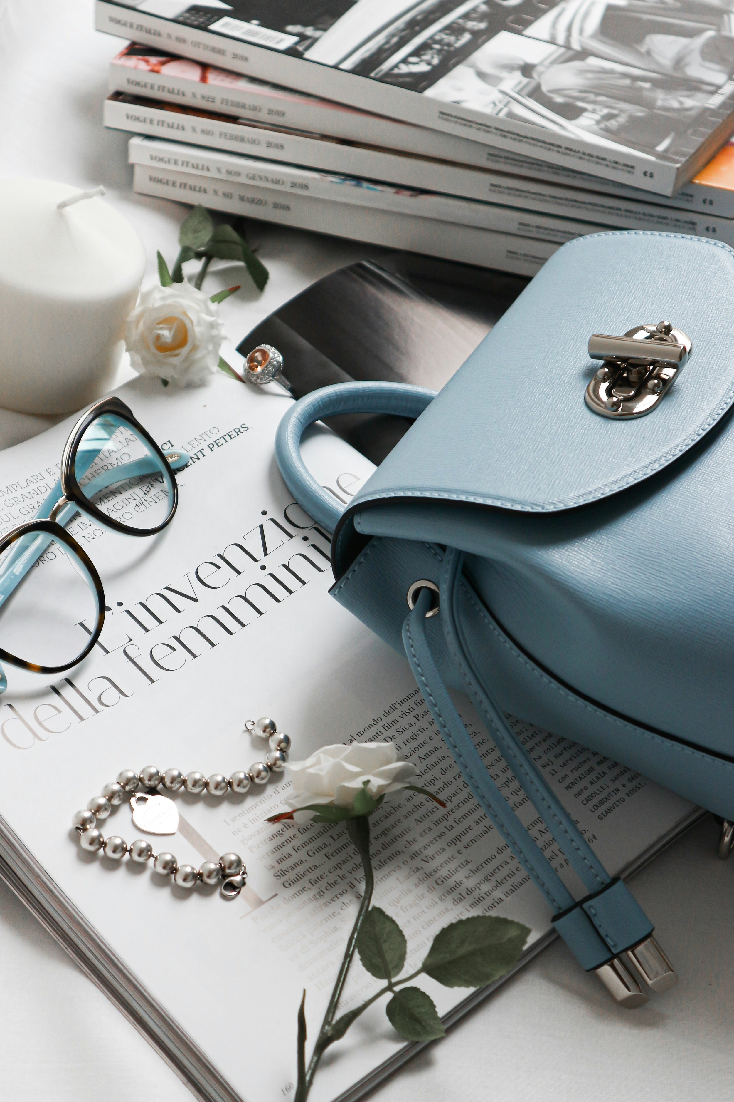 Flat lay of fashion and reading items: glasses, a white candle, white roses, a light blue handbag, a pearl bracelet, corkscrew earrings, a magazine, and a stack of Vogue Italia magazines.