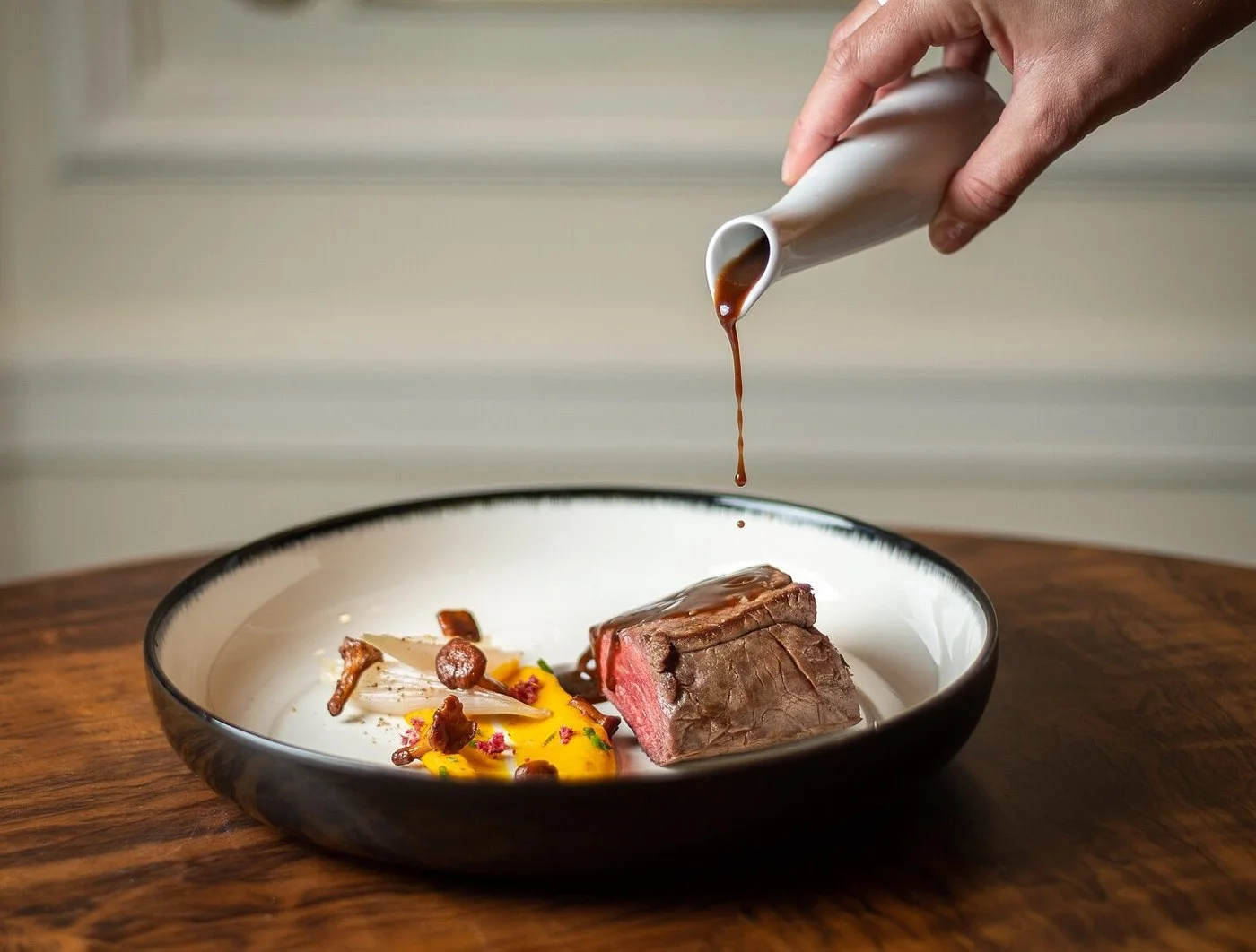A hand pouring brown sauce over a medium-rare steak on a white plate with vegetables, on a wooden table.
