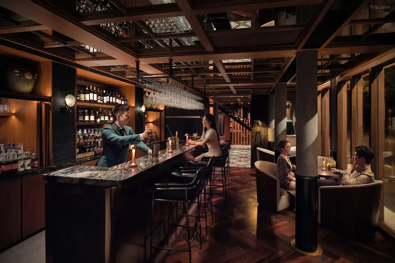 A dimly lit bar with a marble countertop, shelves of liquor bottles, and people sitting at the bar and in cozy booths, with a bartender preparing a drink.