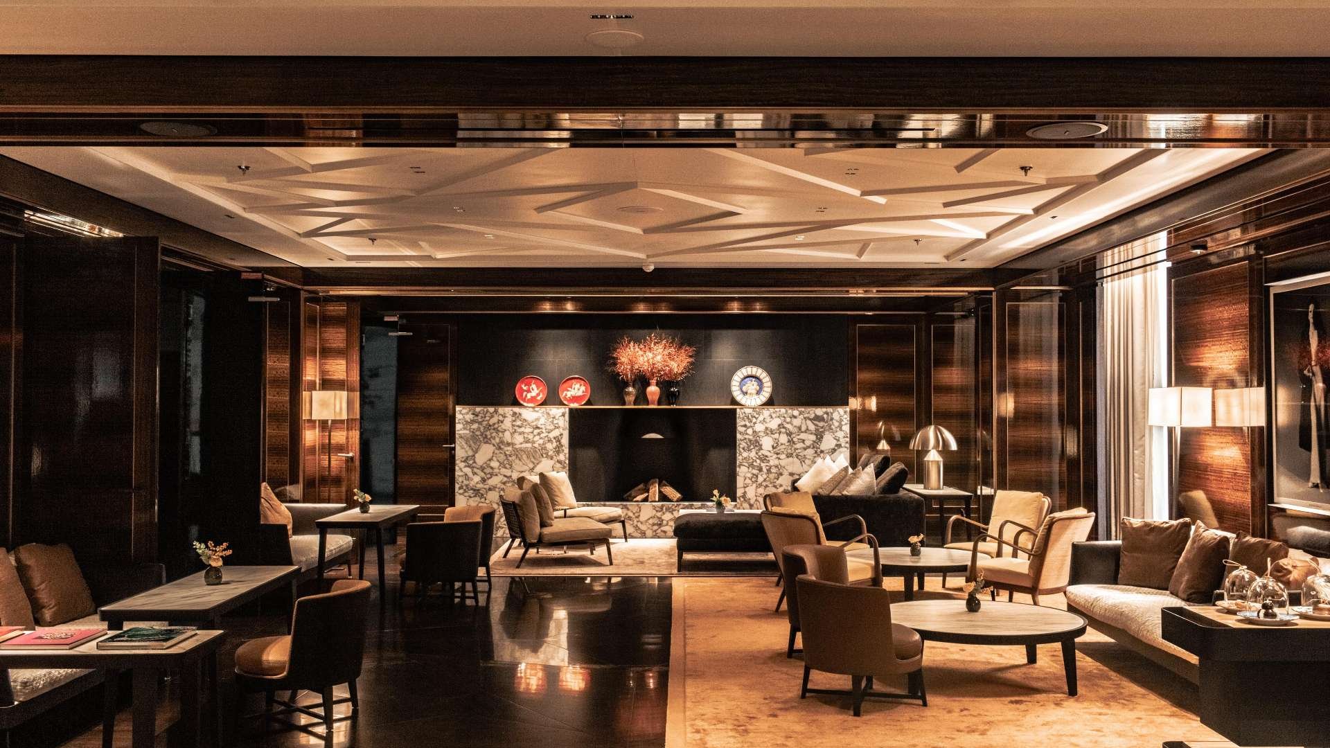 Elegant hotel lobby with dark wood paneling, marble accents, and plush seating arrangements. There is a fireplace with decorative vases and flowers, and warm lighting from lamps and wall fixtures.