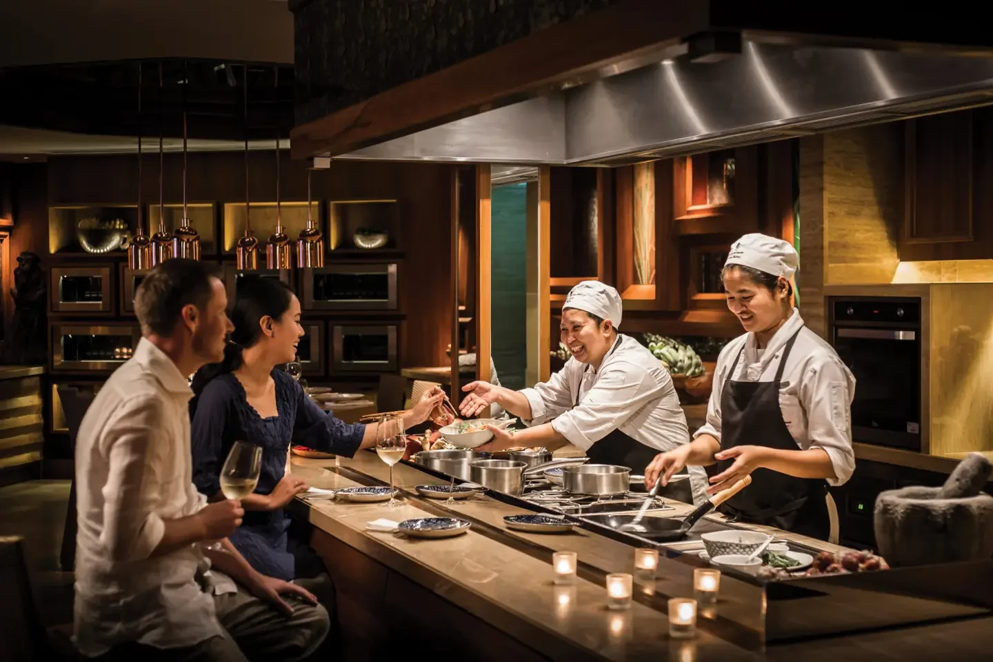 Two chefs serving food to a couple in a restaurant kitchen.