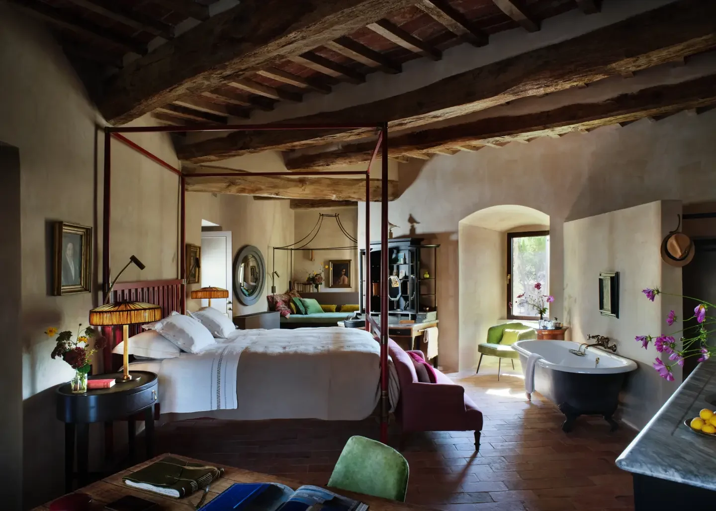 A spacious vintage-style bedroom with a four-poster bed, striped bedding, and wooden beams on the ceiling. The room features a bathtub near a window, colorful chairs, a black shelving unit, and eclectic wall decor including framed paintings and a mir