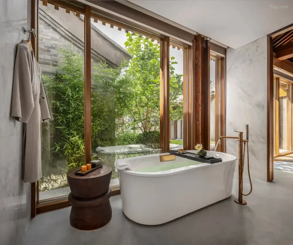 Modern bathroom with a white freestanding bathtub near large floor-to-ceiling windows with wooden frames, overlooking a lush garden with greenery, and a taupe bathrobe hanging on a wall hook.