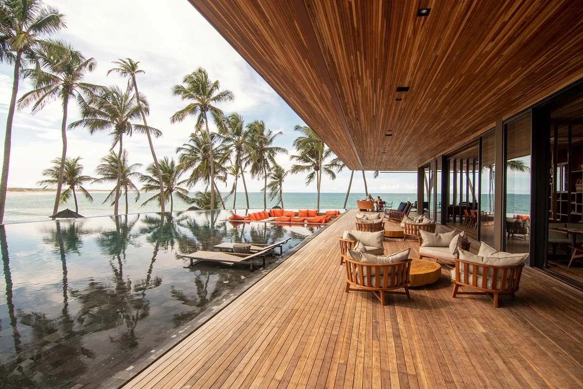 Luxury seaside resort balcony with wooden flooring, outdoor seating with cushions, and palm trees overlooking the ocean.