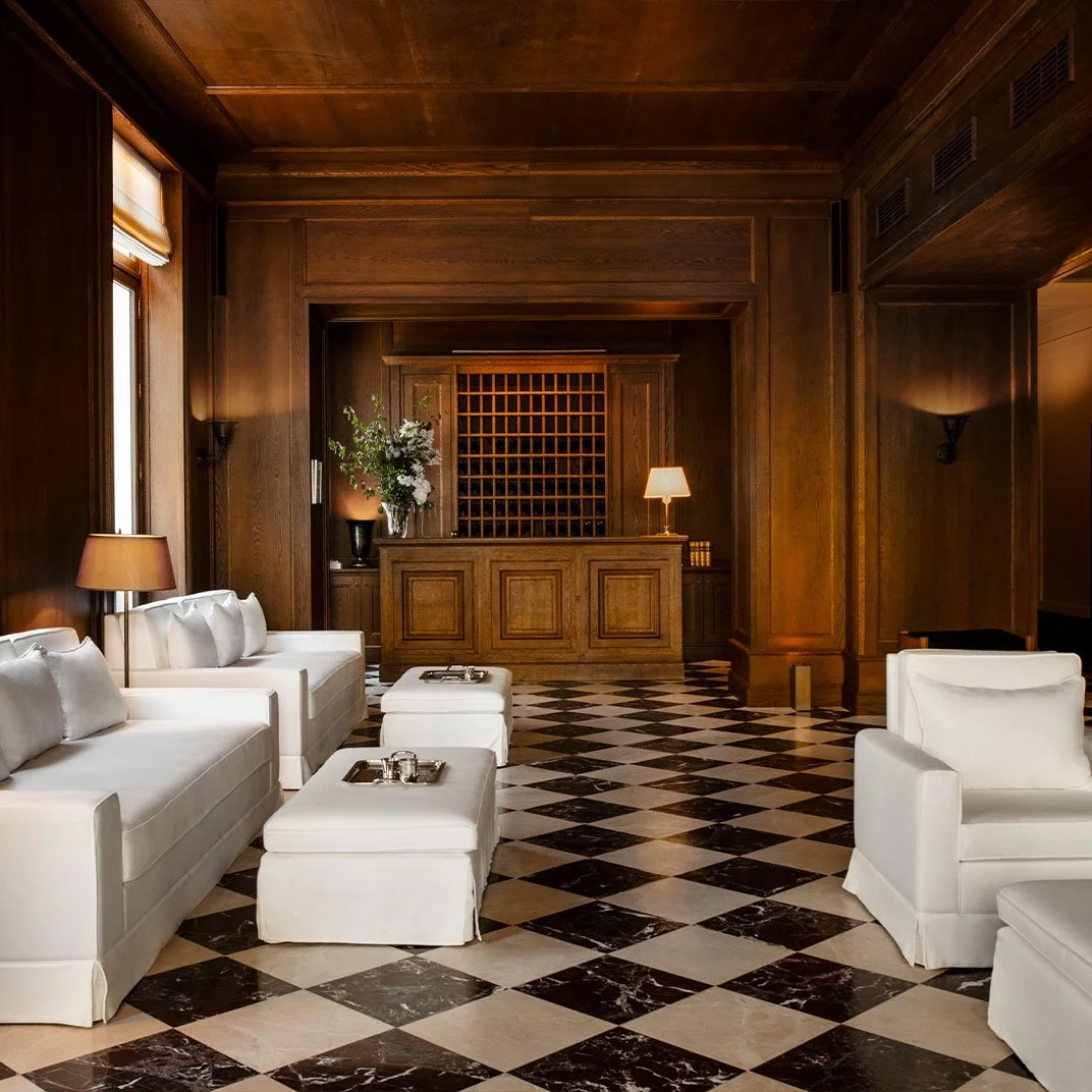 Elegant hotel lobby with dark wood paneling, black and white checkered marble floors, white sofas, and side tables with silver trays, lit by lamps and wall sconces.