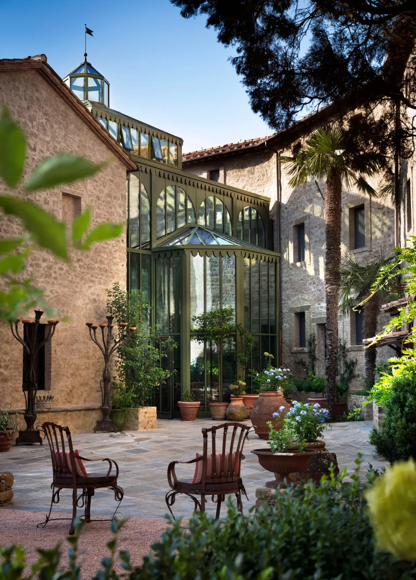 A courtyard with ornate metal chairs, large potted plants, and a glass conservatory attached to old stone buildings, surrounded by lush greenery and tall trees.
