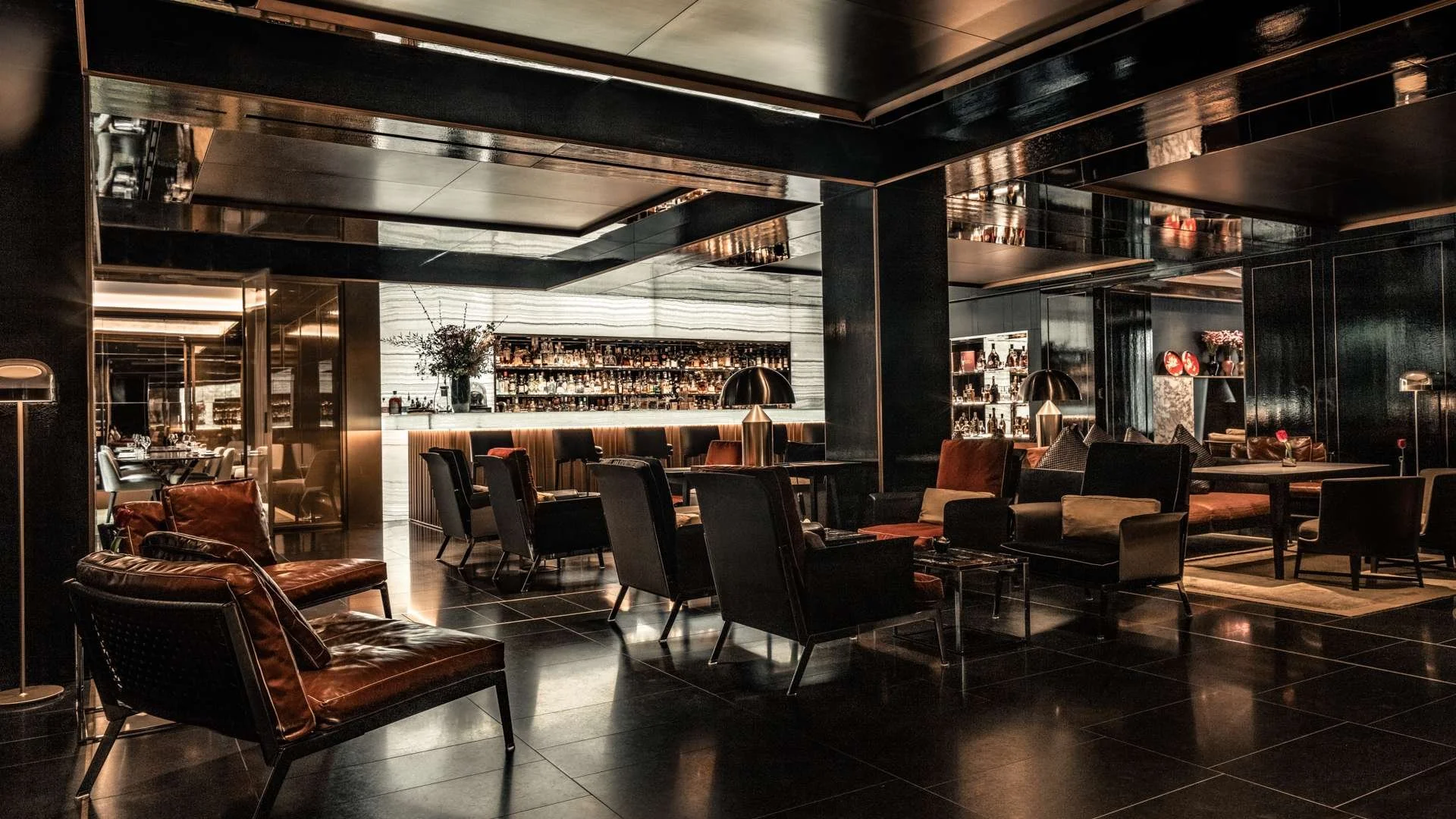 Interior of a luxurious bar and lounge with black and brown leather chairs, a bar with bottles on shelves, and dim lighting creating a cozy atmosphere.
