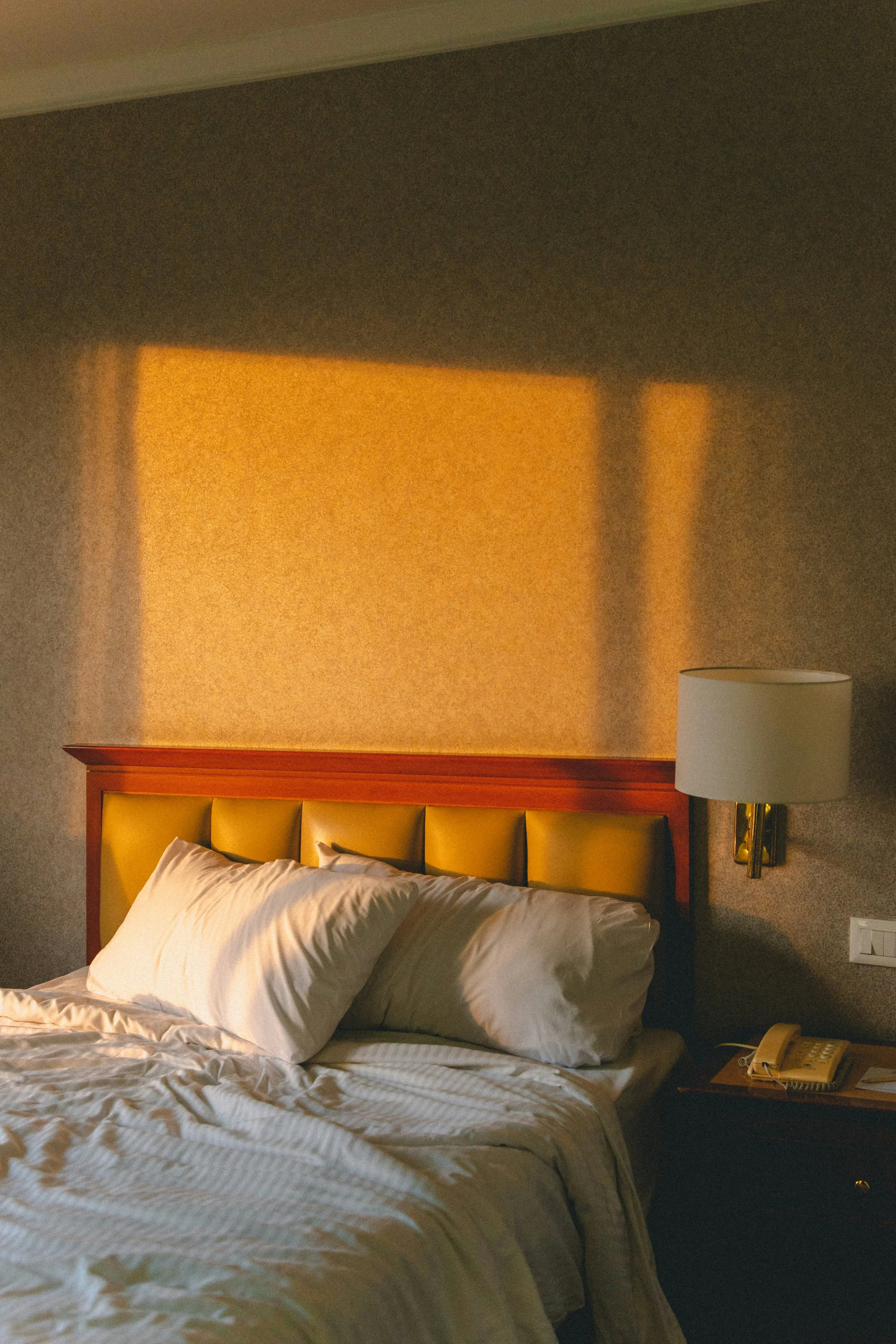 A bedroom with a bed, white pillows, a bedside lamp, and a telephone on nightstand under warm lighting.