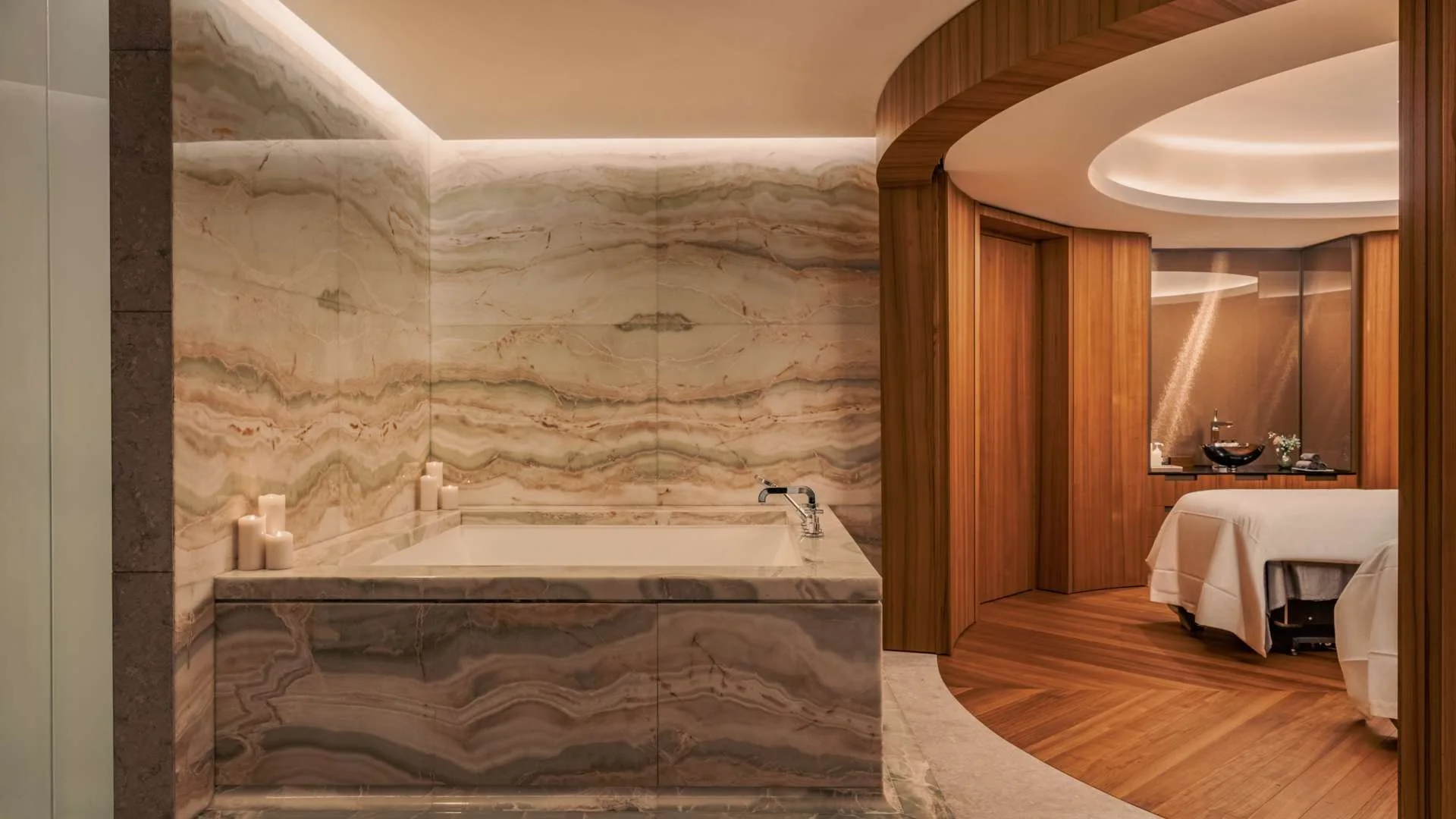 A luxurious bathroom with a built-in bathtub surrounded by marble walls, candles on the edge, and a transitioning wooden-finish room with a bed and decorative washbasin visible in the background.