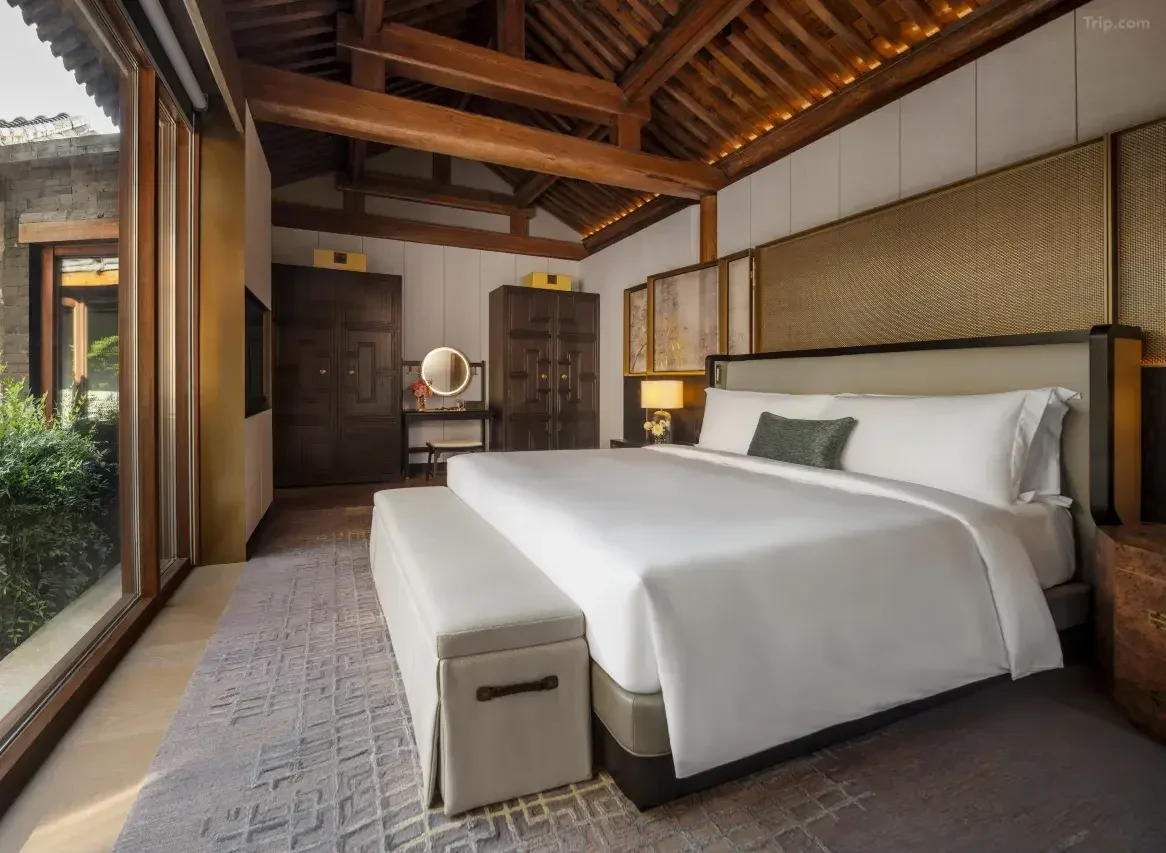 Hotel bedroom with a large bed, bedside lamps, wooden ceiling beams, and a sliding glass door leading outside.