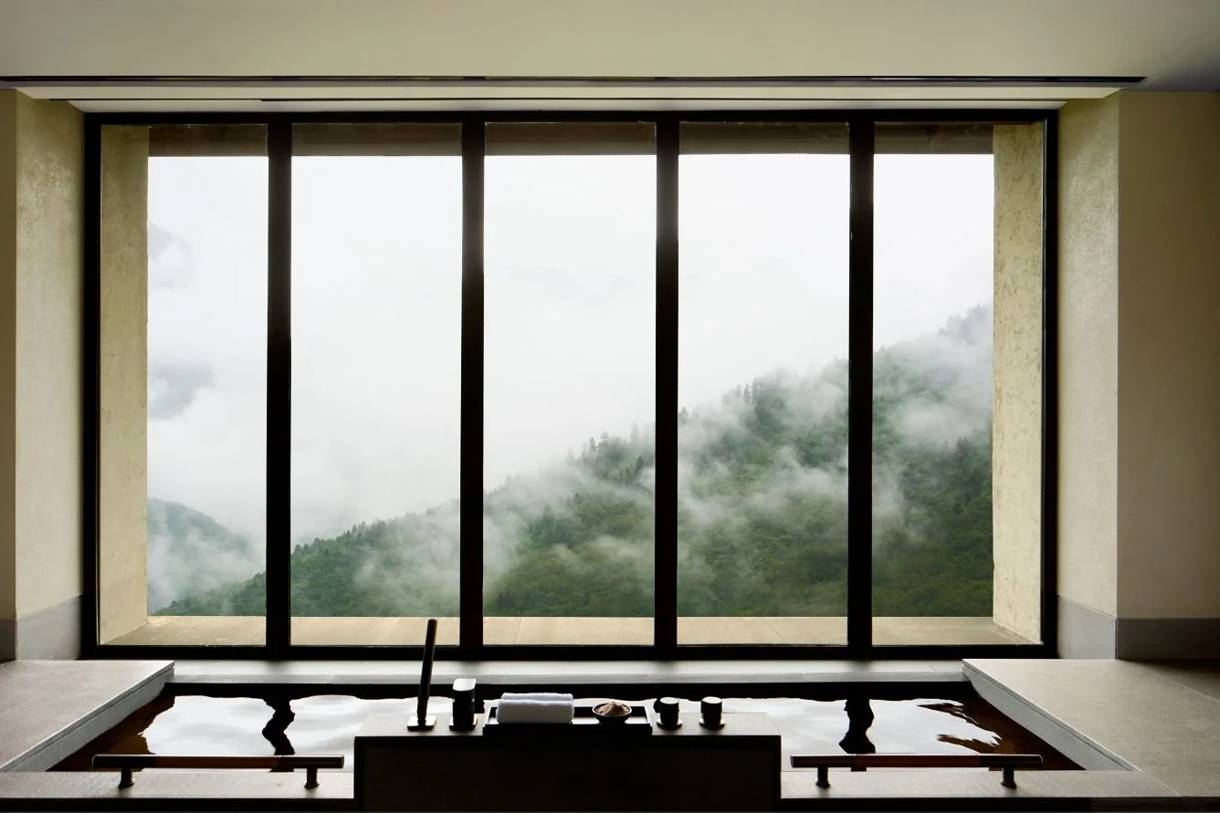 An indoor tub with water and toiletries, overlooking a mountain landscape with clouds and fog through large floor-to-ceiling windows.