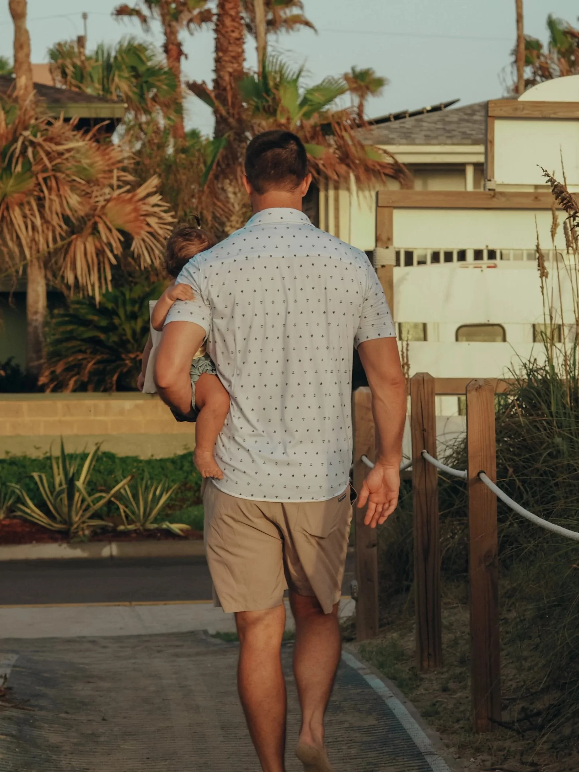A man carrying a child on his shoulder walks along a path near tropical plants and a wooden fence in a residential area.