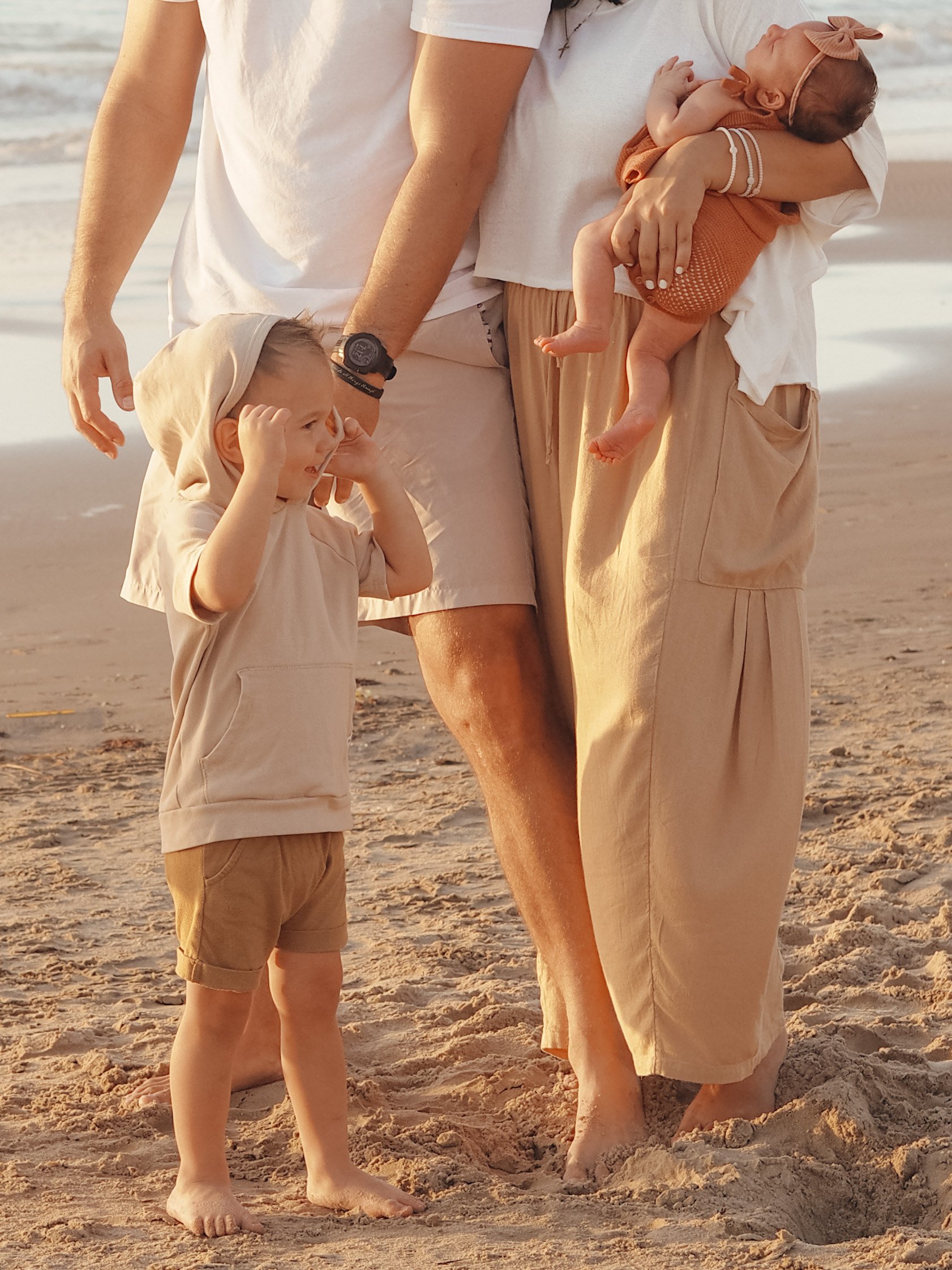 A group of people on the beach, including a woman holding a baby, a young child standing nearby, and an adult with part of the body visible, all dressed in light-colored clothing.