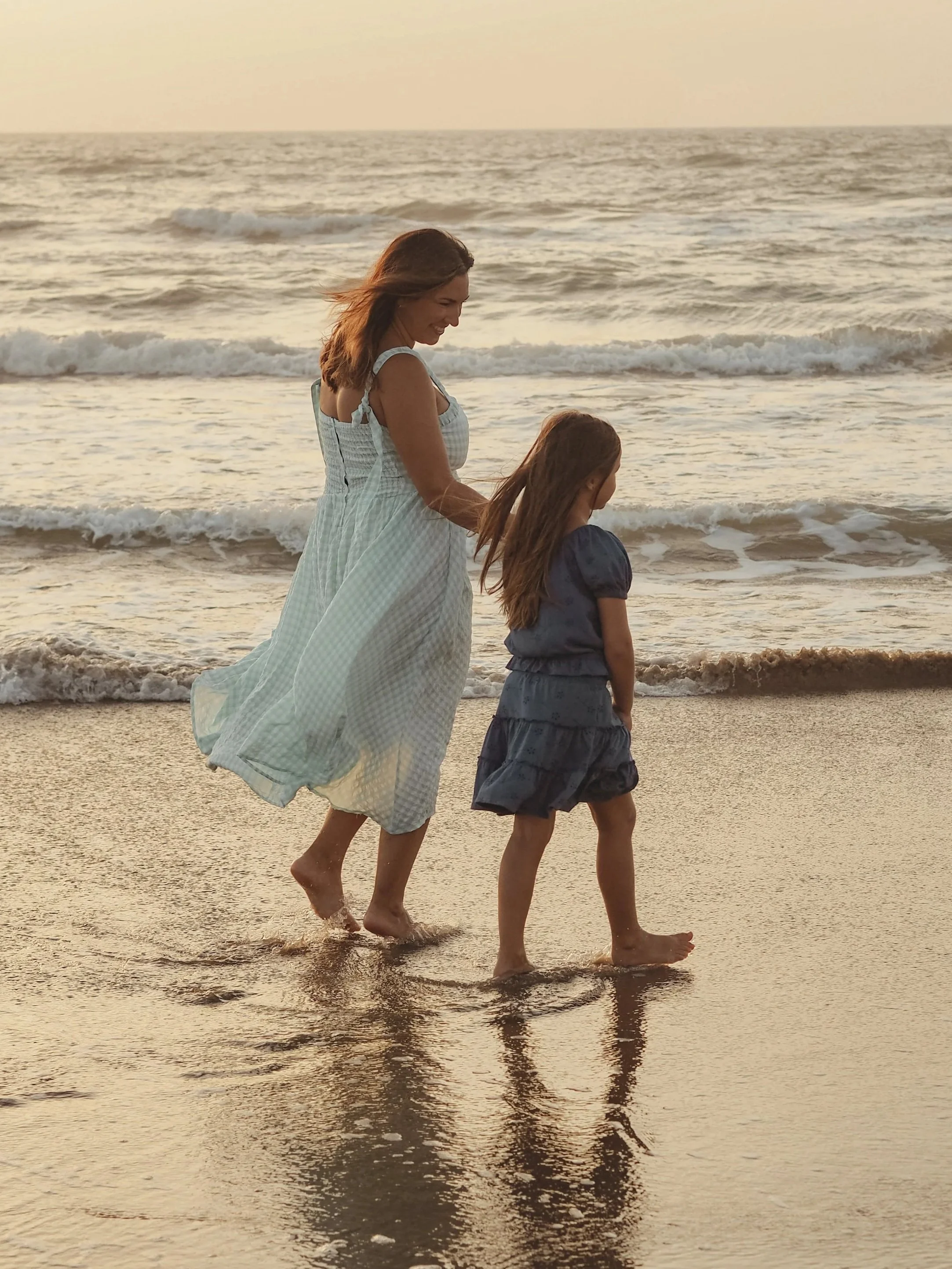 A mother and daughter walking barefoot along the shoreline at sunset, with gentle waves in the background.