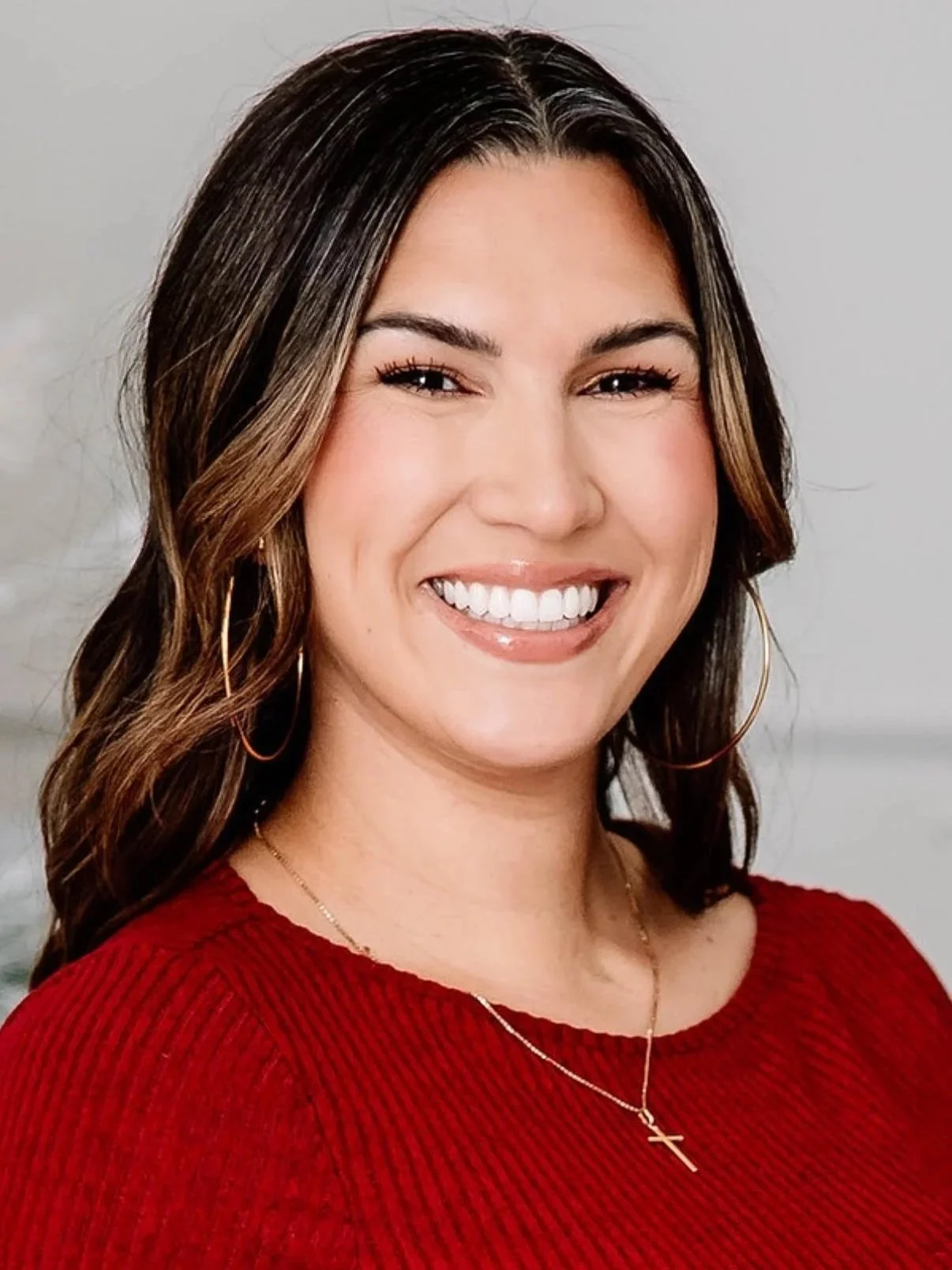Close-up portrait of a smiling woman with shoulder-length brown hair, wearing gold hoop earrings, a red sweater, and a gold cross necklace, against a neutral background.