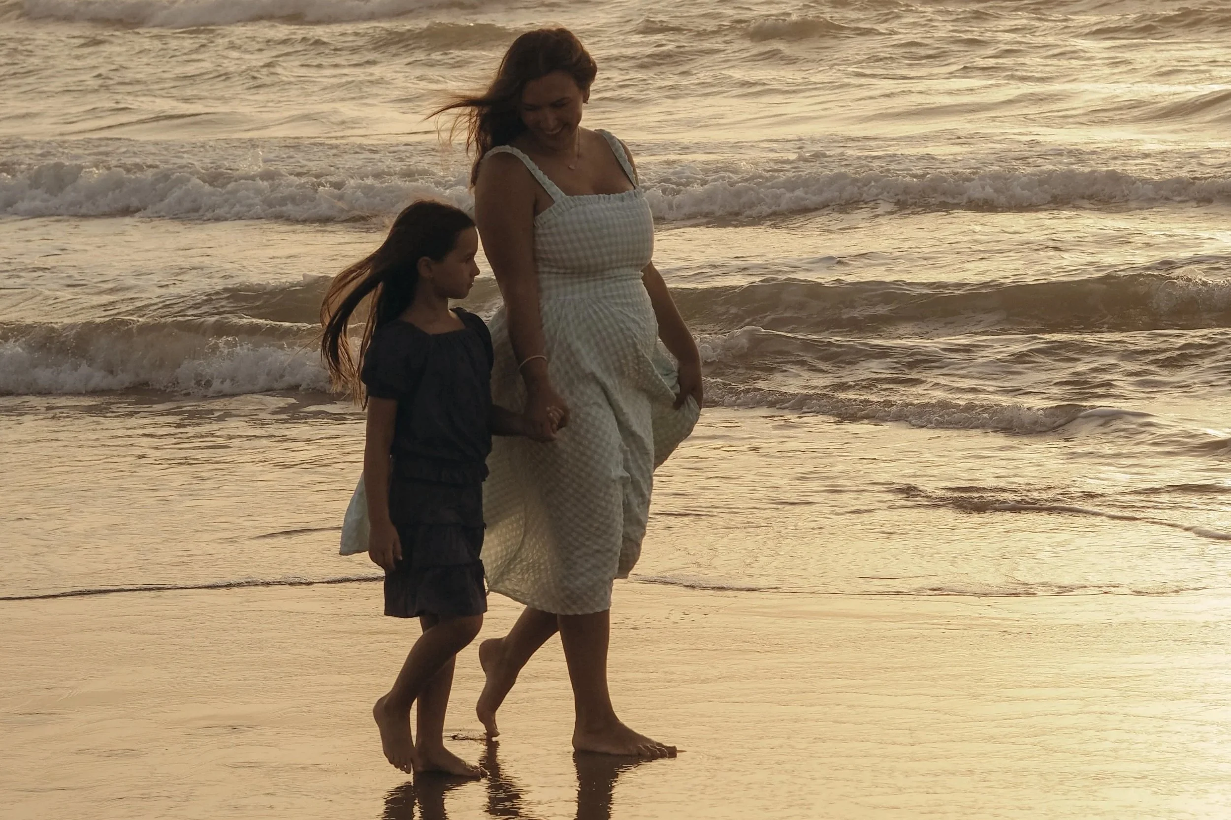 A woman and young girl walking along the beach at sunset, holding hands, with gentle waves in the background.