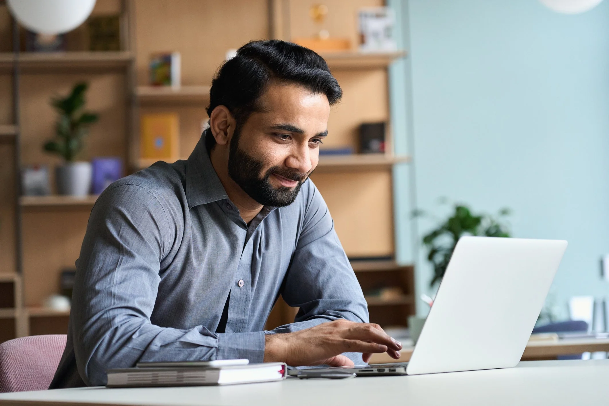 A man with dark hair and a beard working on a laptop at a desk in an office or home office space.